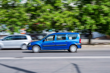 Car rides on city road with ghost effect. Buildings, green trees and parked cars on background.