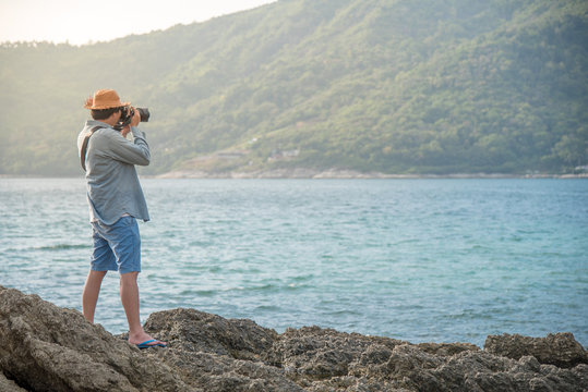 young Asian man photographer on the rock taking photos of tropical island during sunset, seascape for summer holiday and vacation travel concepts