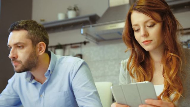 Caucasian young pretty woman with red hair siting in kitchen with her husband using tablet, smiling. Indoor.