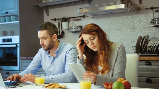 Serious young couple siting in kitchen. Young beautiful woman using digital tablet while her husband using laptop. Indoor.