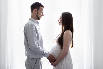 Fototapeta premium family, pregnancy and parenthood concept - happy pregnant couple in front of the window