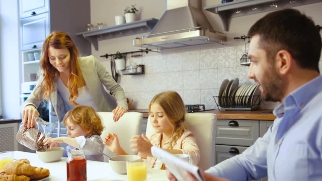Caucasian little boy, girl and father siting on the table in kitchen. Father reading newspaper, mother filling flakes in plate of son, dauther eating flakes with milk.