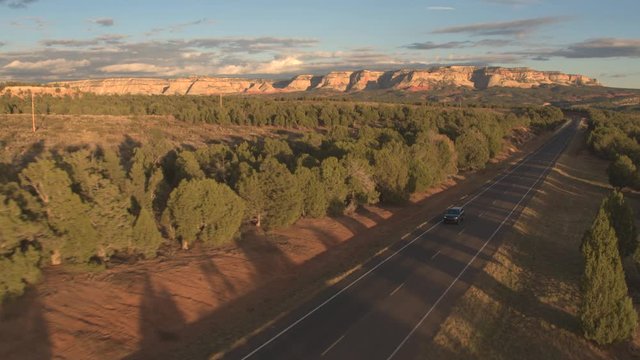 AERIAL: Black SUV car driving along the empty road through pine forest valley with mesa mountains in the background. Jeep on a journey across United States traveling through Utah on sunny evening