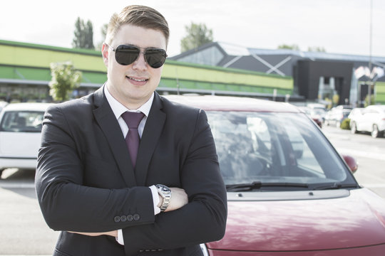 Young ambitious businessman hand crossed looking at the camera while standing in front of his car. Suit and tie businessman in front of a modern office building.