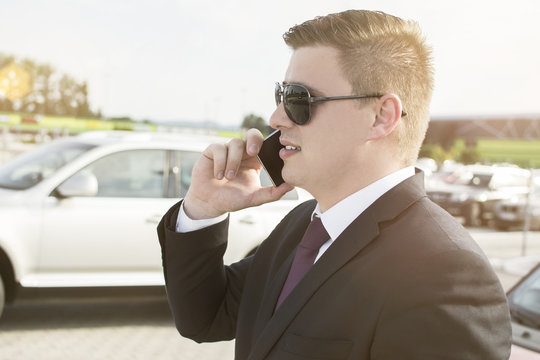 Close-up of a businessman in front of his office building. Young ambitious businessman talking on his phone while standing in a car parking lot. Suit and tie businessman having a conversation.
