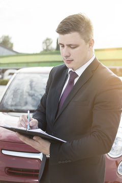 Closeup of a man filing a paper form on a clipboard. Modern businessman standing in a parking lot in front of the car. Lens Flare in the background.
