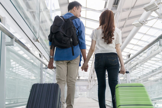 Young happy asian couple form back view  carrying suitcase luggage in airport terminal. Couple holding hand and traveling abroad together, Air travel or holiday vacation concept