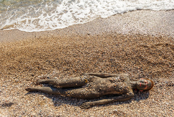 Beautiful woman laying on the beach and sea waves splash near her.Top view. Woman smearing mud mask on body, Spa outdoor and cosmetics. Recreation tourism and lifestyle, body care, natural therapy.