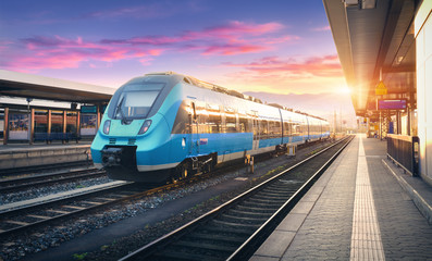 Moderni brzi prigradski vlak na željezničkoj stanici i šareno nebo s oblacima za zalaska sunca u Europi. Industrijski krajolik s plavim putničkim vlakom na željezničkoj platformi. Pozadina željeznice