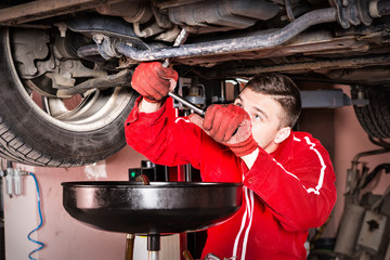 Male car mechanic in uniform working underneath a lifted car and changing motor oil