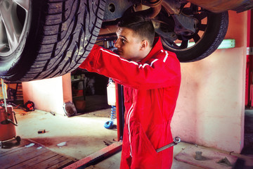 Young car mechanic in uniform is repairing wheel while working underneath a lifted car