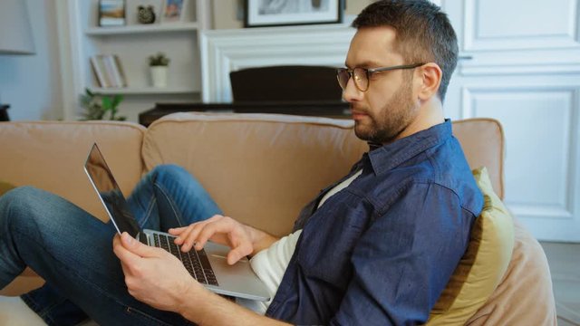 Attractive caucasian man in glasses siting on the couch and typing on laptop on home background. Indoor.