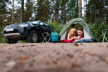 Beautiful couple lying in a tent, camping on the background of fire. Next to folding chairs, camping a mug of warm soup and a Hiking backpack. Beside their off-road machine.