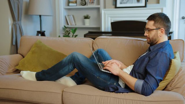 Concentrated caucasian man in glasses working in home, siting on the sofa and typing on laptop. Indoor.