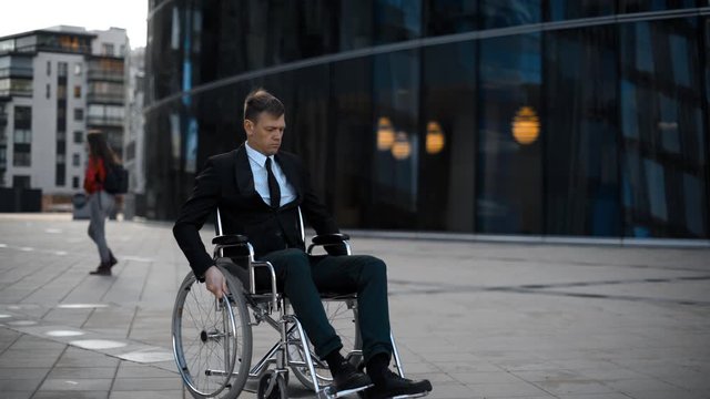 Handsome invalid man rolling in wheelchair and looking depressed on You. Portrait in 4k UHD near businesscenter outdoor glass teal and orange. Failure concept.