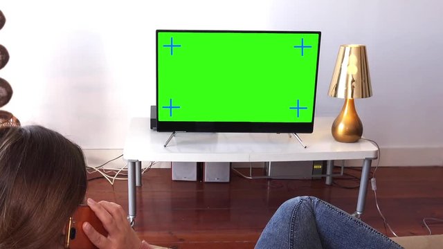 Girl Receives Phone Call While Watching TV Lying On Couch. Young woman talks on the phone while watching television green screen. Shot behind models shoulders