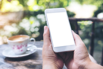 Mockup image of hands holding white mobile phone with blank screen on vintage wooden table and nature background