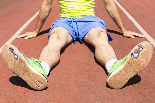 An exhausted athlete on a running track wearing broken green running shoes with big holes in the sole.