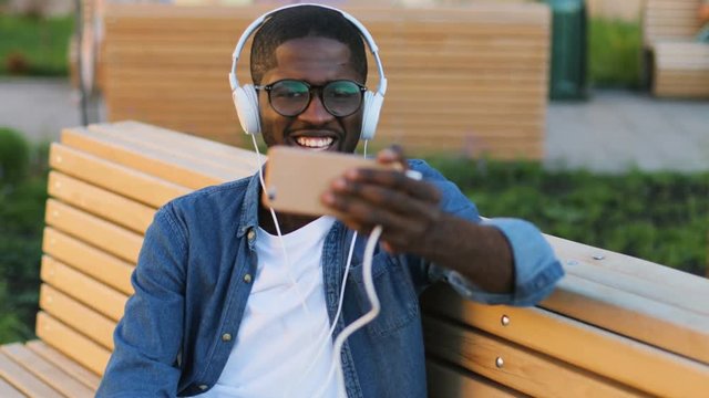 Attractive african man in glasses with earphones using smart phone for video chatting with friend siting on the bench in city.