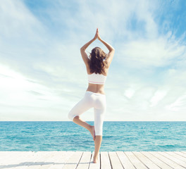 Attractive, beautiful and young girl in white sportswear meditating on a wooden pier at summer. Yoga, sport, recreation, vacation and traveling concept.
