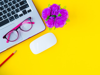 feminine desk workspace with office accessories including  mouse laptop, glasses, red pencil, note book and pink flower on yellow background.