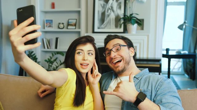 Young attractive couple in love taking selfie on smart phone camera, making funny faces and smiling at home.