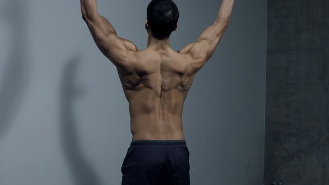 A male fitness model displays his back muscles for the camera. Filmed in an isolated medium shot.