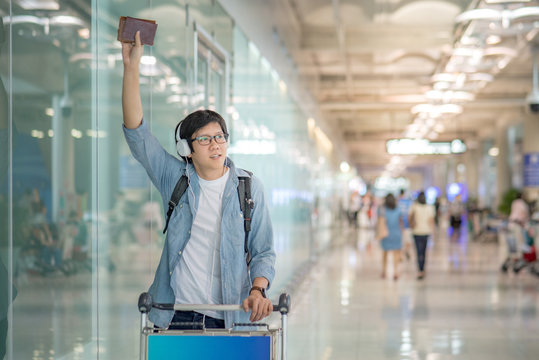 Young Asian Man With Airport Trolley Waving His Hand In The International Airport Terminal, Arrival From Travel Abroad