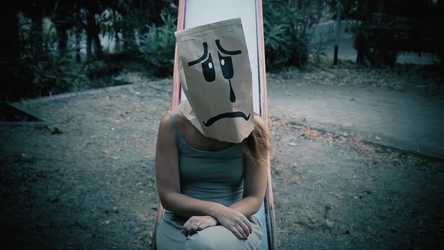 A breadbag face at a public park, sitting on a slide, with a sad depressed expression. Handheld shot.
