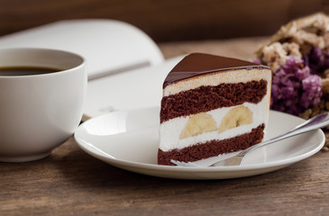 Banana chocolate cake on white plate served with black coffee. Chocolate cake layered with whipped cream and banana look so delicious. Enjoy eating at cafe restaurant with chocolate cake and coffee.