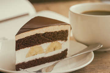 Banana chocolate cake on white plate served with black coffee. Chocolate cake layered with whipped cream and banana look so delicious. Enjoy eating at cafe restaurant with chocolate cake and coffee.