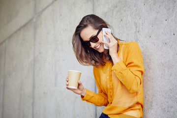Beautiful young lady talking on phone and drinking coffee while outside in the city