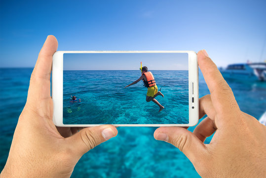 Hand holding a smart phone taking photograph boys jumping into the sea.
