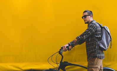 Fototapeta na wymiar Handsome Adult Man In Sunglasses Walks With A Bicycle Along A Yellow Wall Copy Space