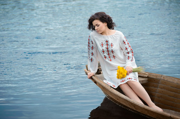 Portrait of a beautiful girl in Slavic clothes. Young woman with a bouquet of dandelions is sitting in a wooden boat. Summertime