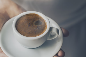 Closeup of tasty coffee espresso with tasty foam in small ceramic cup. Male hands holding warm hot drink.