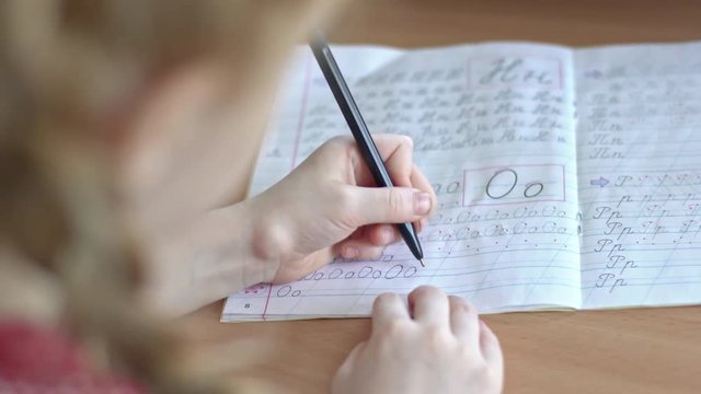 Over the shoulder shot with PAN of unrecognizable little girl with braided hair practicing writing letters in notebook
