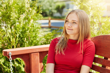 Outdoor portrait of pretty female with straight fair hair and pleasant appearance wearing red sweater sitting at bench near green plants looking aside having much dreams. Summer vacation concept