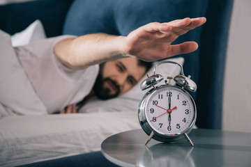 Bearded young man trying to turn off alarm clock while lying in bed at morning