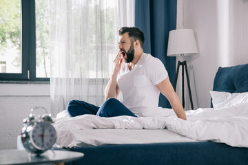 Bearded young man in pajamas yawning while sitting on bed in morning