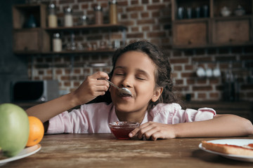 portrait of little girl with eyes closed eating sweet jam from teaspoon