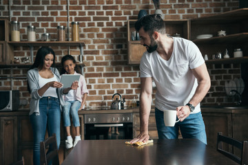 father drinking coffee and cleaning table while mother and daughter using digital tablet at kitchen