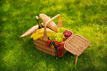Wicker picnic basket with fresh fruits, wine bottle, baguette and plaid on green meadow at daytime