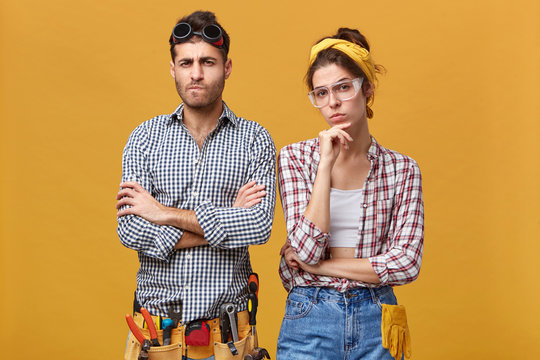 Handsome young electrician wearing belt kit with plier, flexible ruler, wrench, screwdriver and hammer folding arms, standing next to his female colleague, both having skeptic and distrustful looks