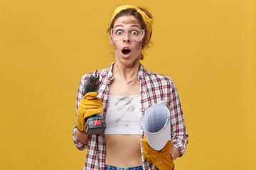 Portrait of surprised woman wearing protective goggles, checkered shirt and white top holding drill...