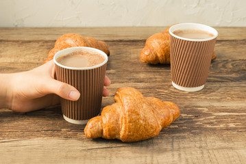 Tasty fresh croissants with coffee in a paper cup on old wooden background. A girl holding a glass of coffee in hand.
