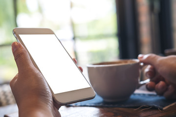 Mockup image of hand holding white mobile phone with blank screen and coffee cup on wooden table in cafe