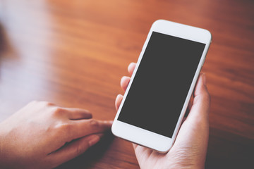 Mockup image of hands holding white mobile phone with blank black screen on vintage wooden table