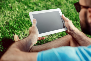 Close-up of casual dressed young man using modern digital tablet while sitting at the park, sun light