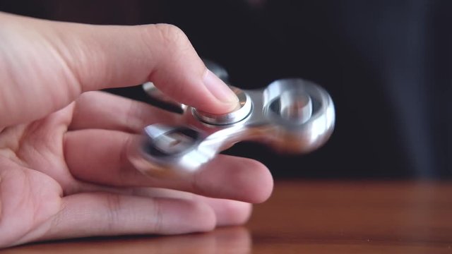 A hand holding and playing metal silver color fidget spinner with people using mobile phone in background